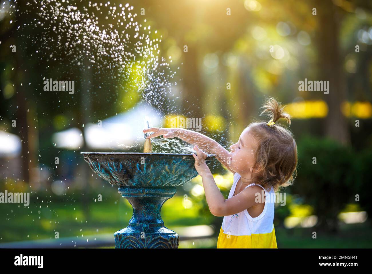 Ein lustiges kleines Mädchen spielt mit dem Sprühnebel eines Trinkwasserbrunnens im Park. Kindheit, Kind, Sommerzeit, Hitze. Solarschutz für Chil Stockfoto