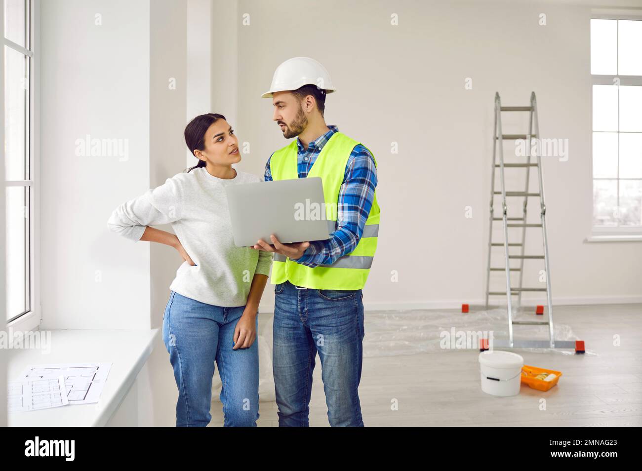 Eine attraktive junge Frau spricht mit einem Baumeister in einem weißen Helm, der im Wohnzimmer auf einem Laptop die Reihenfolge der Reparaturen vorführt. Stockfoto