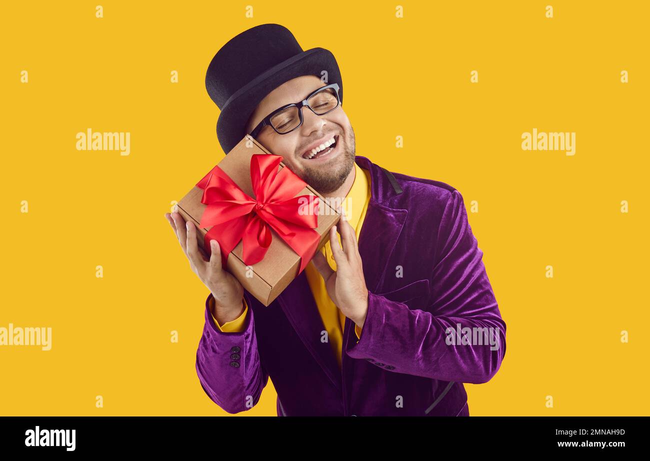 Ein lustiger Mann in exzentrischer Kleidung mit befriedigendem Gesichtsausdruck wird gegen eine Geschenkbox gekuschelt. Stockfoto