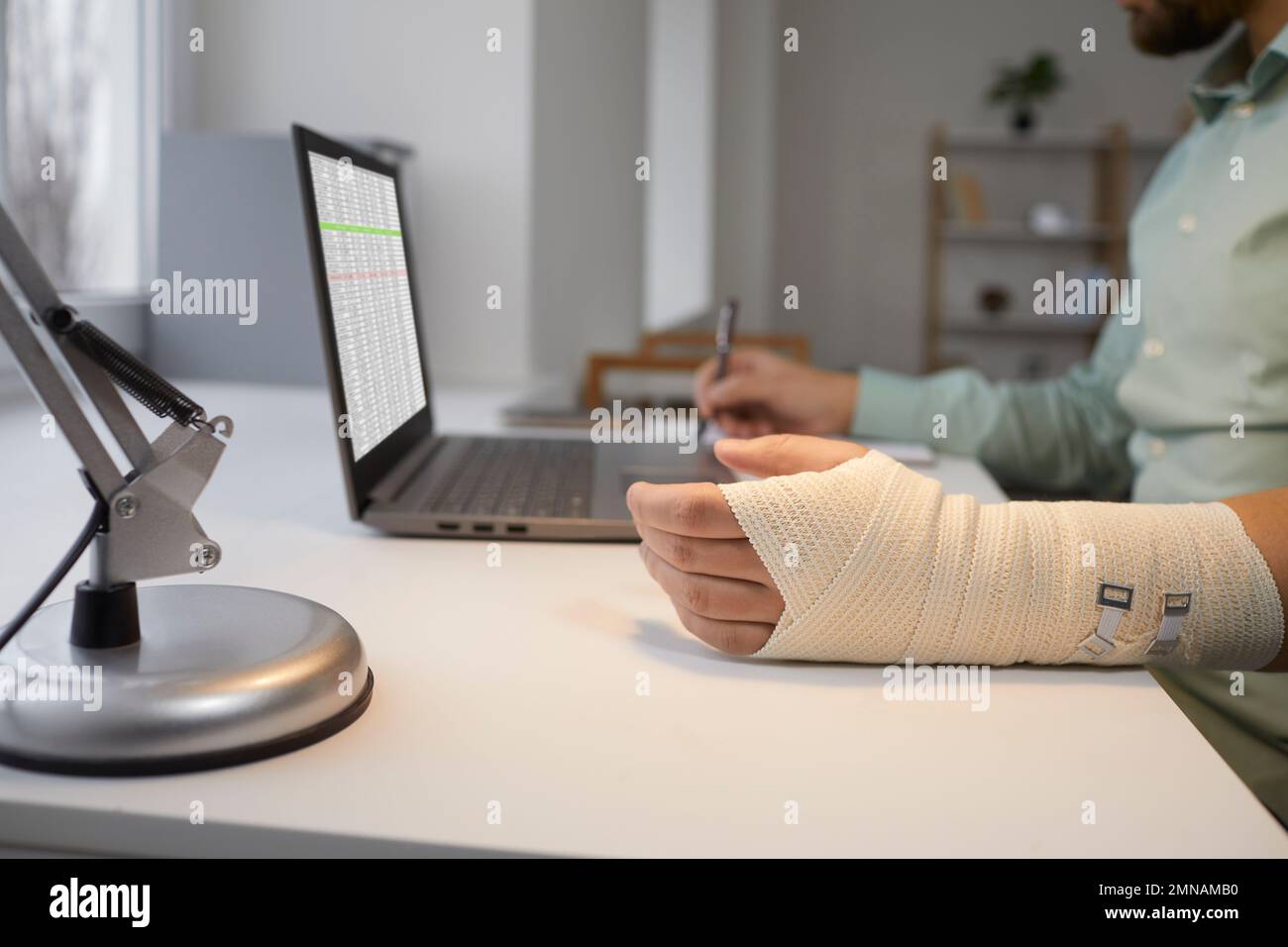Ein bärtiger Mann mit gebrochenem Arm arbeitet an einem Laptop, sitzt an seinem Schreibtisch und macht sich Notizen in einem Notizbuch für Berichte. Stockfoto