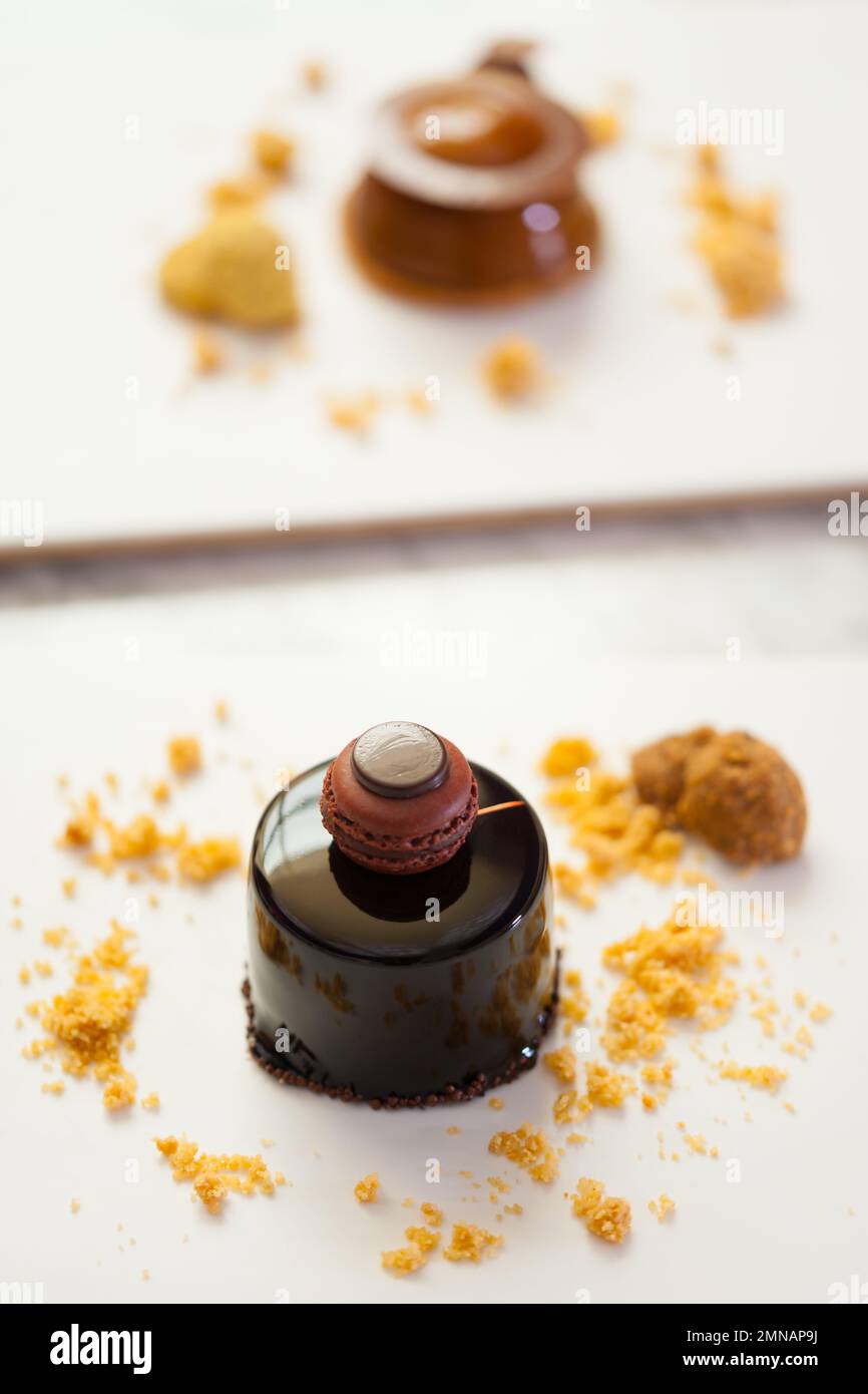 Nahaufnahme von Mini-Mousse-Kuchen aus dunkler Schokolade mit französischem Makkaron, Keksen und Mandelkrümeln auf dem Teller. Moderne Dessertpanzerung im Hintergrund. Stockfoto