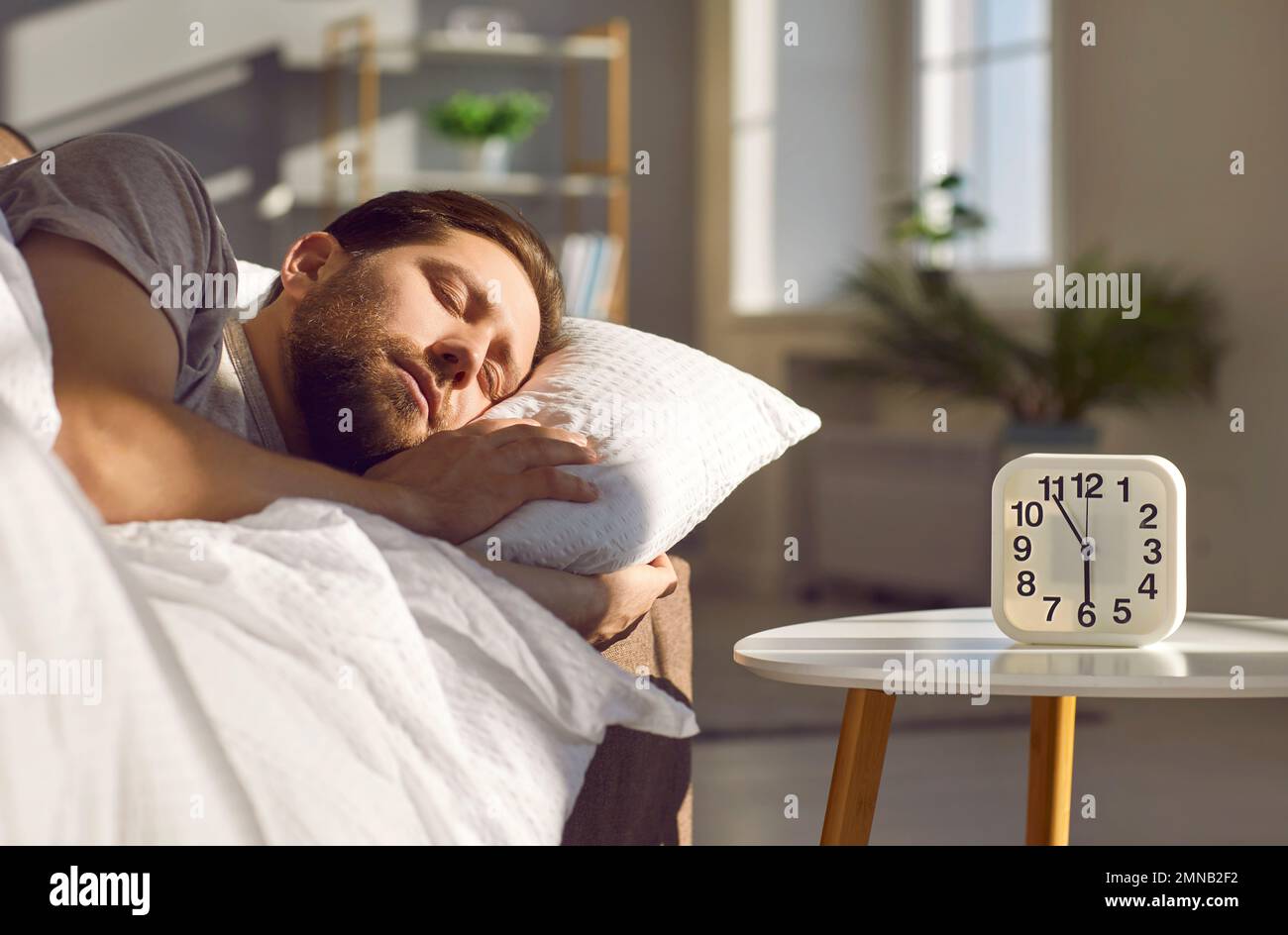 Ein gutaussehender Mann schläft in seinem Schlafzimmer mit Wecker auf seinem Nachttisch. Stockfoto