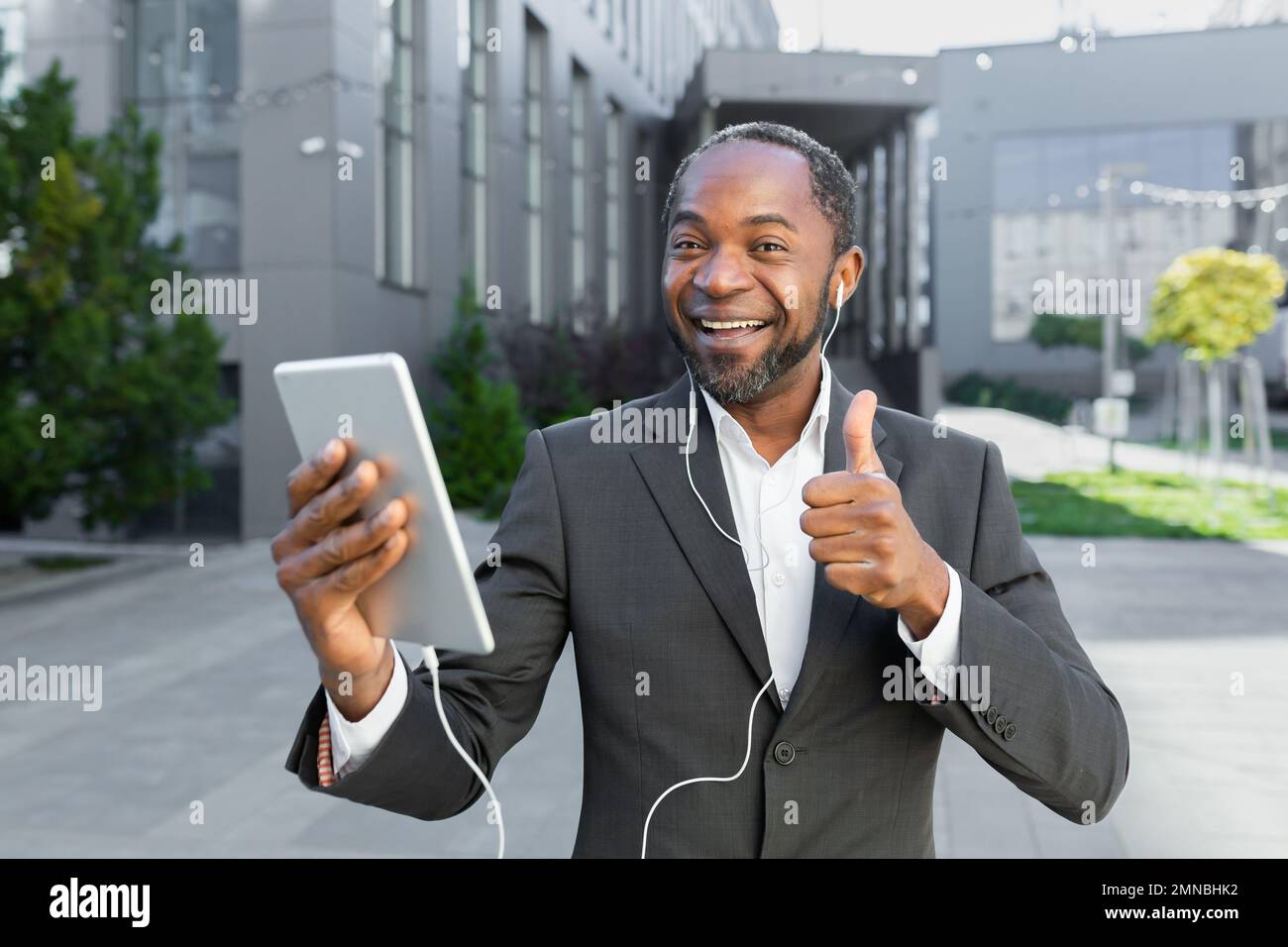Ein junger afroamerikanischer Mann steht draußen in der Nähe eines Bürozentrums, trägt Kopfhörer und spricht bei einem Videoanruf. Hält ein Tablet in seinen Händen, zeigt einen Superhelden, lächelt, schaut in die Kamera. Stockfoto