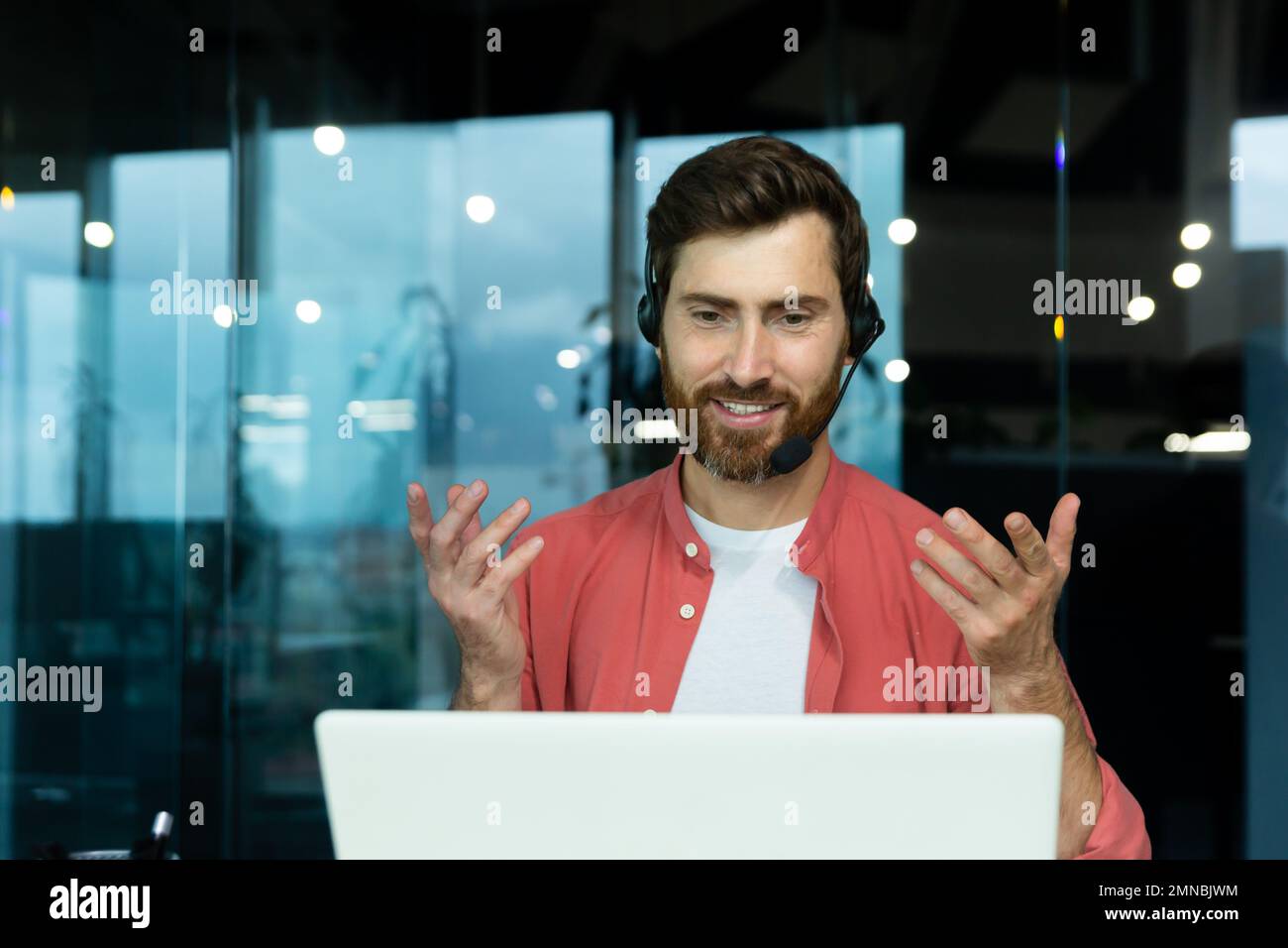 Fernunterricht. Porträt eines jungen Lehrers in einem Headset in einem roten Hemd sitzt im Büro am Tisch, arbeitet online an einem Laptop und unterrichtet durch einen Videoanruf. Das erklärt er mit seinen Händen, lächelt Stockfoto