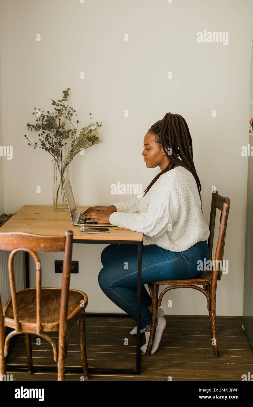 Fröhliche afroamerikanische Frau, die ihren Laptop benutzt, während sie im Wohnzimmer auf einem Stuhl sitzt - Student, Videoanruf und Arbeit zu Hause - Konzept Stockfoto
