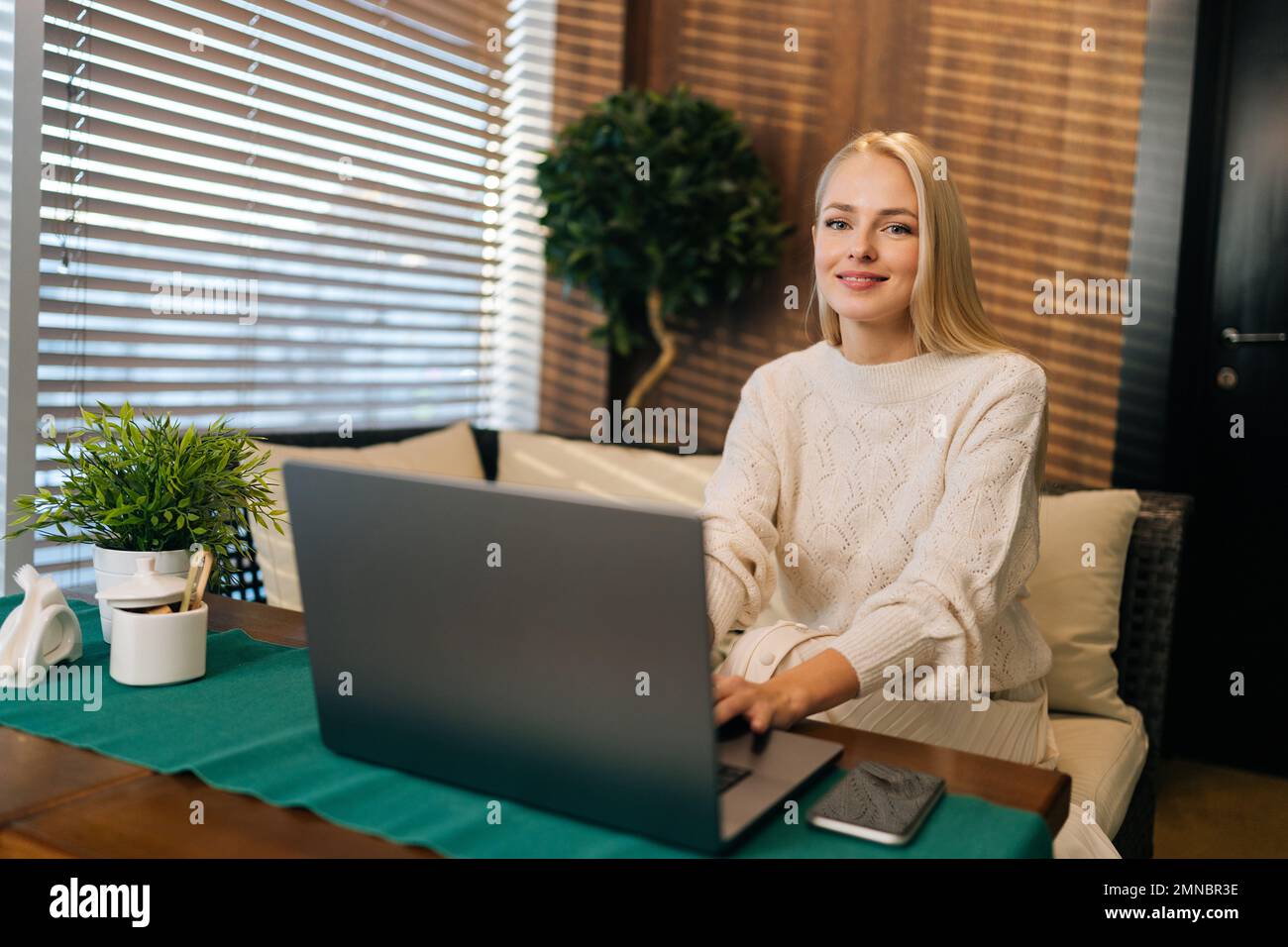Porträt einer charmanten jungen Frau mit langen blonden Haaren, die am Tisch im Café sitzt, während sie einen Laptop benutzt und in die Kamera lächelt. Stockfoto