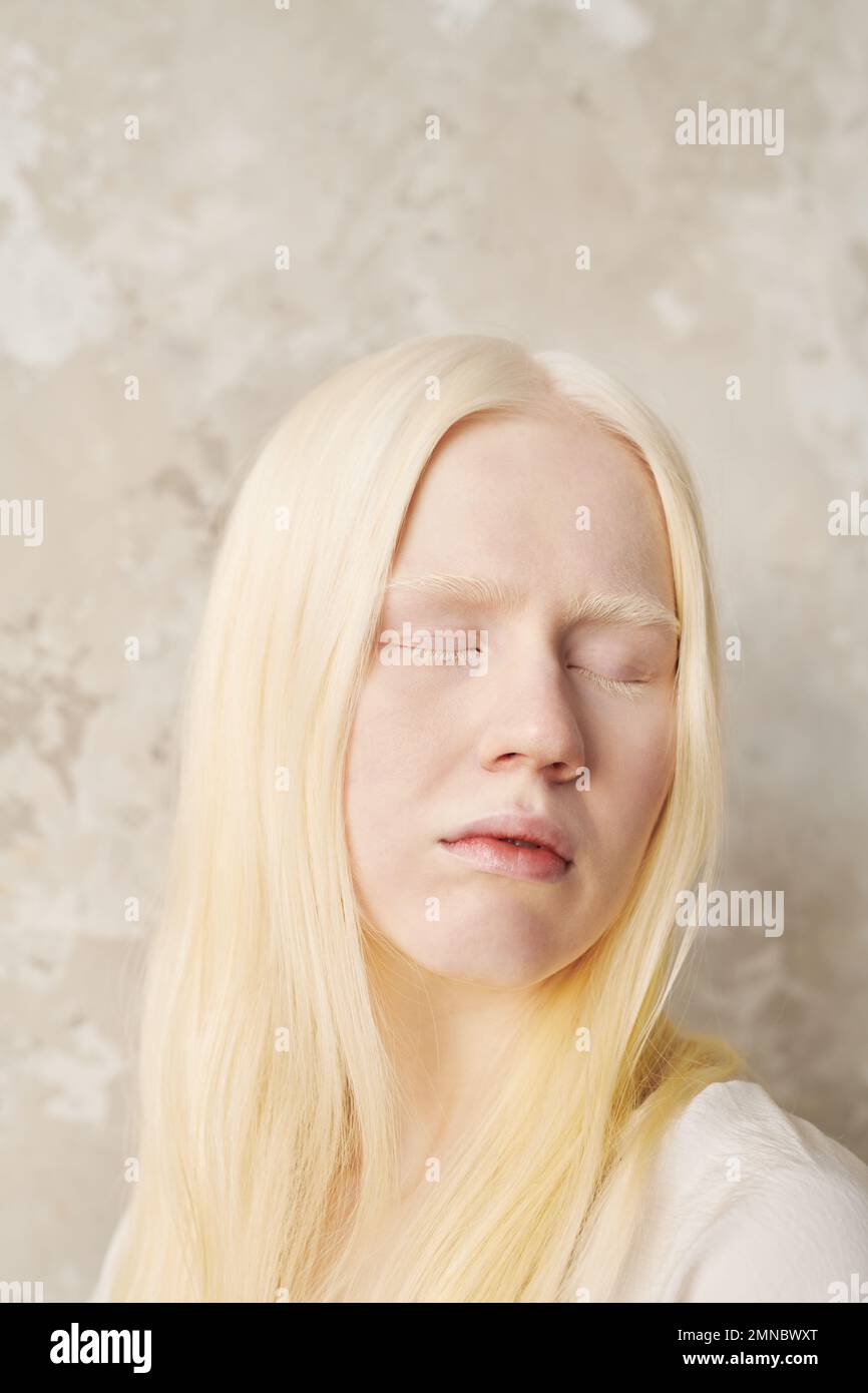 Junge Albino-Frau mit blasser Haut und sehr schönen Haaren, die ihre Augen geschlossen hält, während sie vor der Kamera gegen die weiße Wand steht Stockfoto