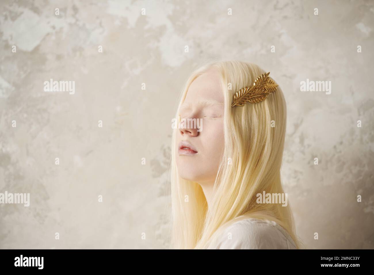 Junge, wunderschöne Albino-Frau mit goldener Haarnadel in langen blonden Haaren, die ihre Augen geschlossen hält, während sie gegen die Marmorwand steht Stockfoto