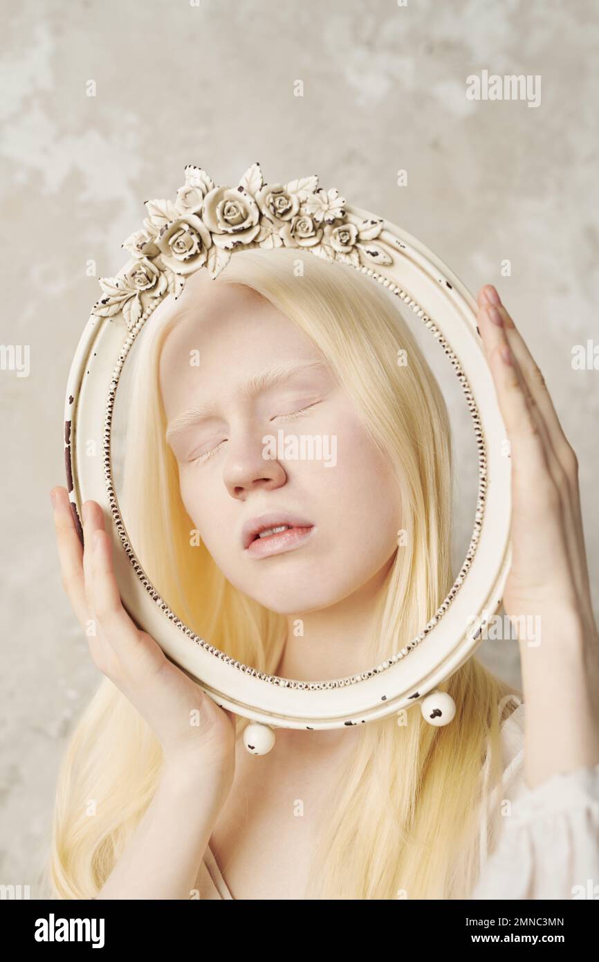 Junge Albino-Frau hält die Augen geschlossen, während sie während der Fotosession im Studio einen dekorativen ovalen Rahmen vor ihr Gesicht hält Stockfoto