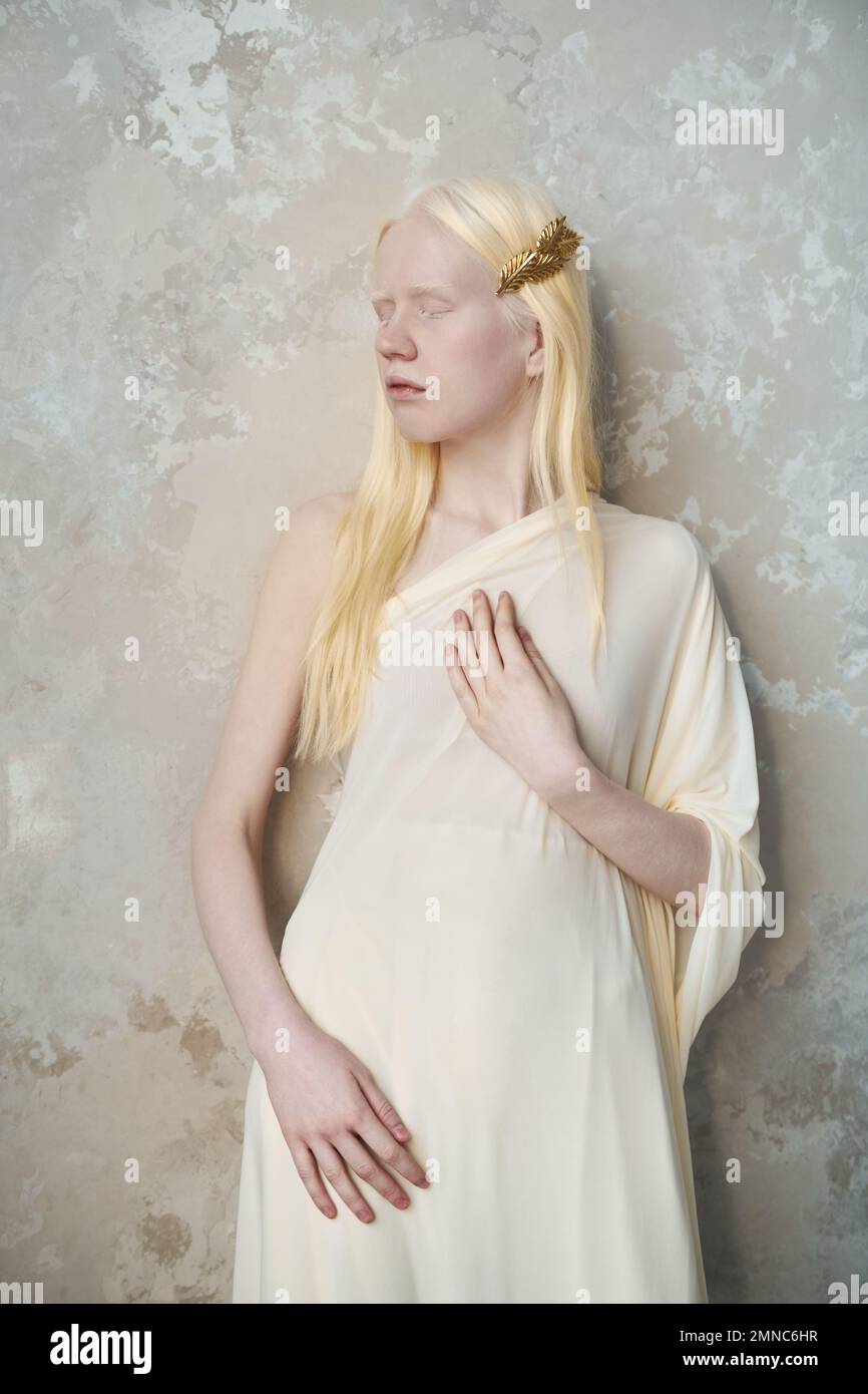 Junge, ruhige Albino-Frau in weißer Kleidung, die ihre Augen geschlossen hält, während sie während der Fotosession an der Marmorwand vor der Kamera steht Stockfoto