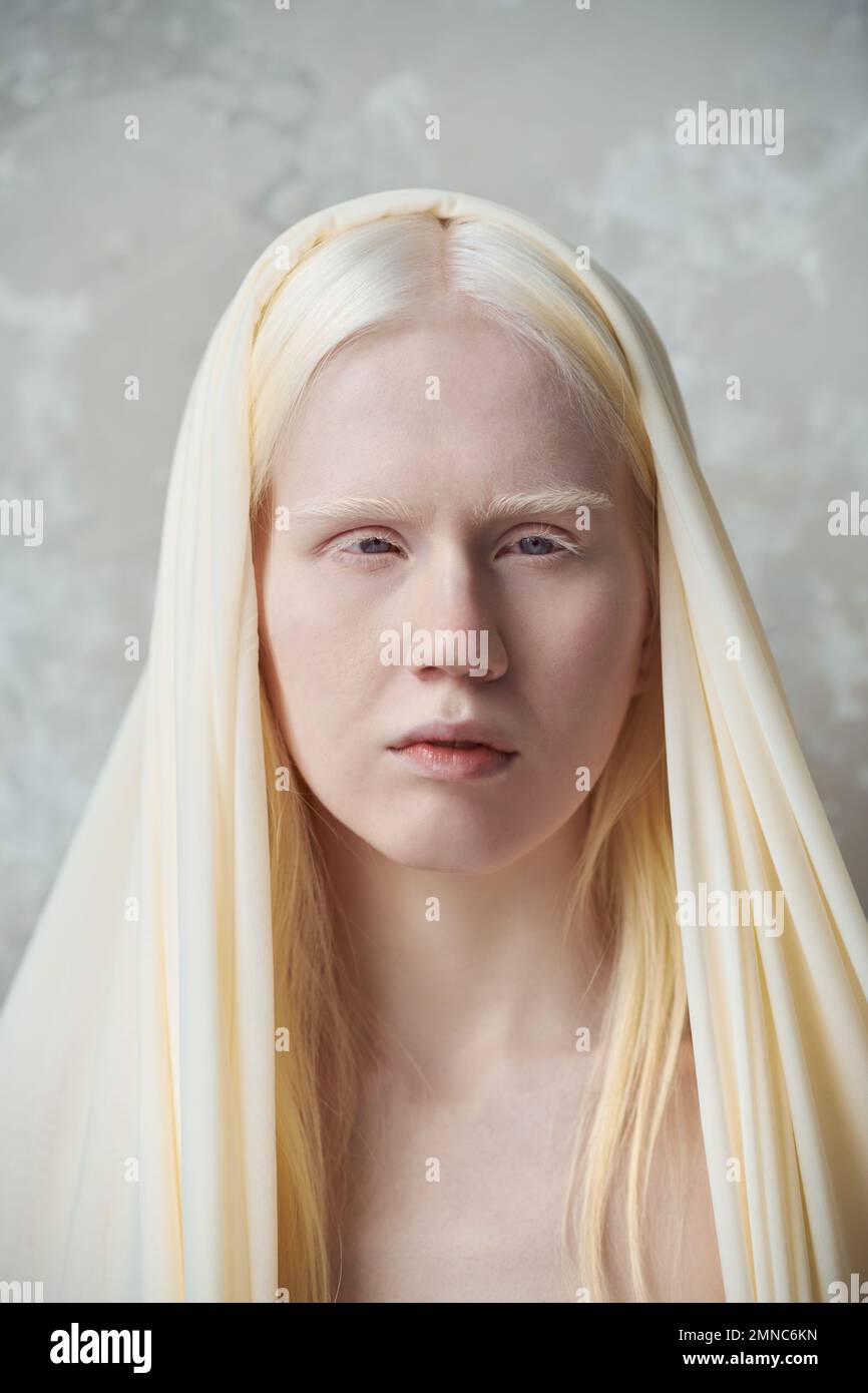Junge, ruhige Albino-Frau mit weißer Abdeckung auf dem Kopf, die während einer Fotosession im Studio vor der Kamera gegen die Marmorwand steht Stockfoto