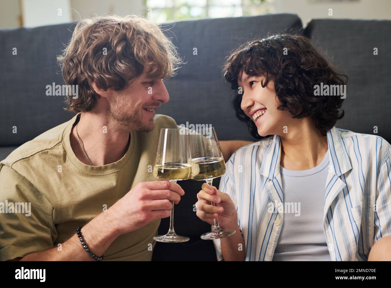 Junger, fröhlicher Ehemann und Ehefrau, die mit Champagnerflöten klingeln und sich mit einem Lächeln anschauen, während sie sich zu Hause auf der Couch entspannen Stockfoto