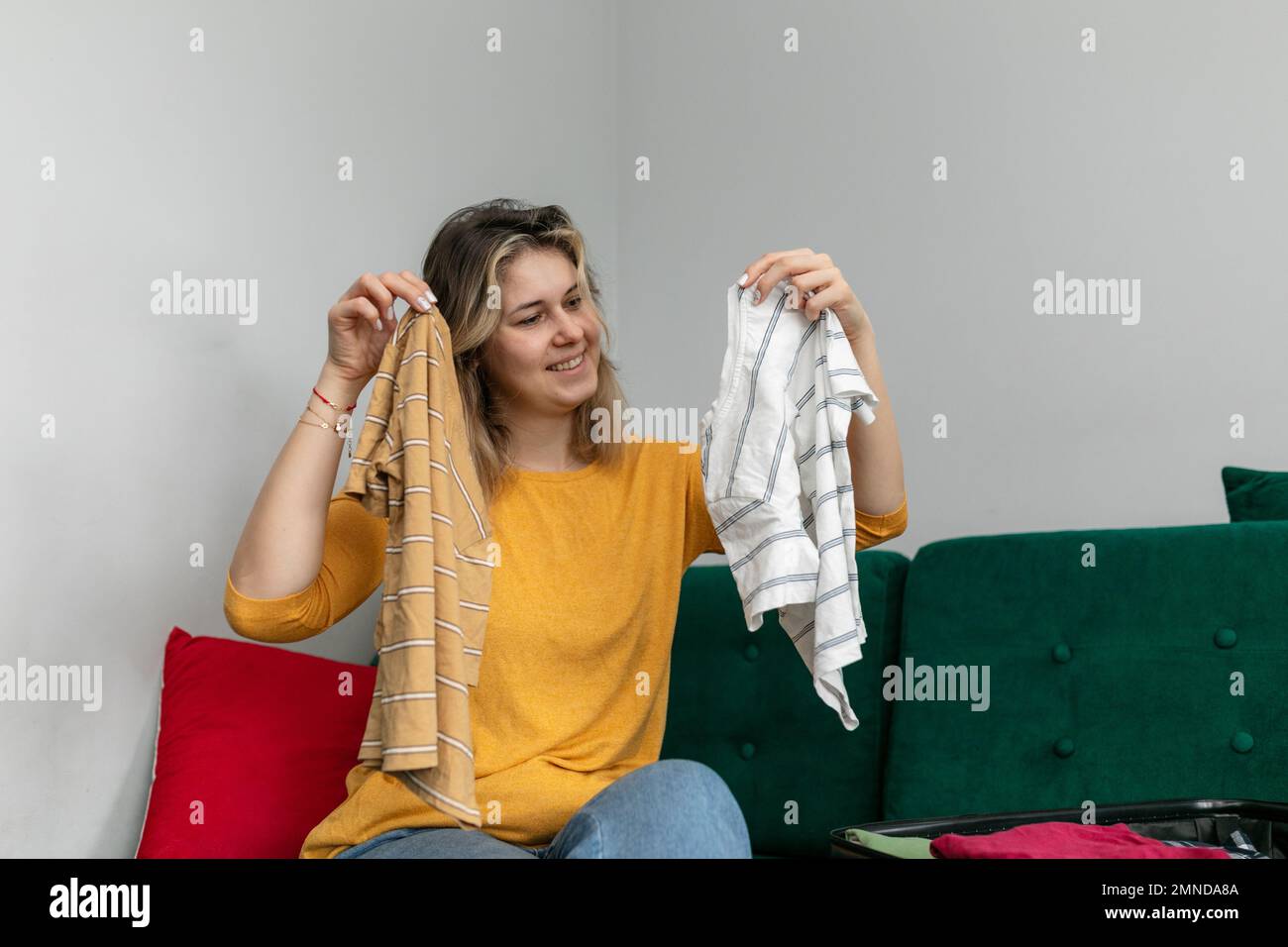 Eine lächelnde Frau hält gestreifte T-Shirts in den Händen, die zu Hause auf dem Sofa sitzen. Alternative Wahl. Klappen Sie die Kleidung in Ihr Gepäck. Stockfoto