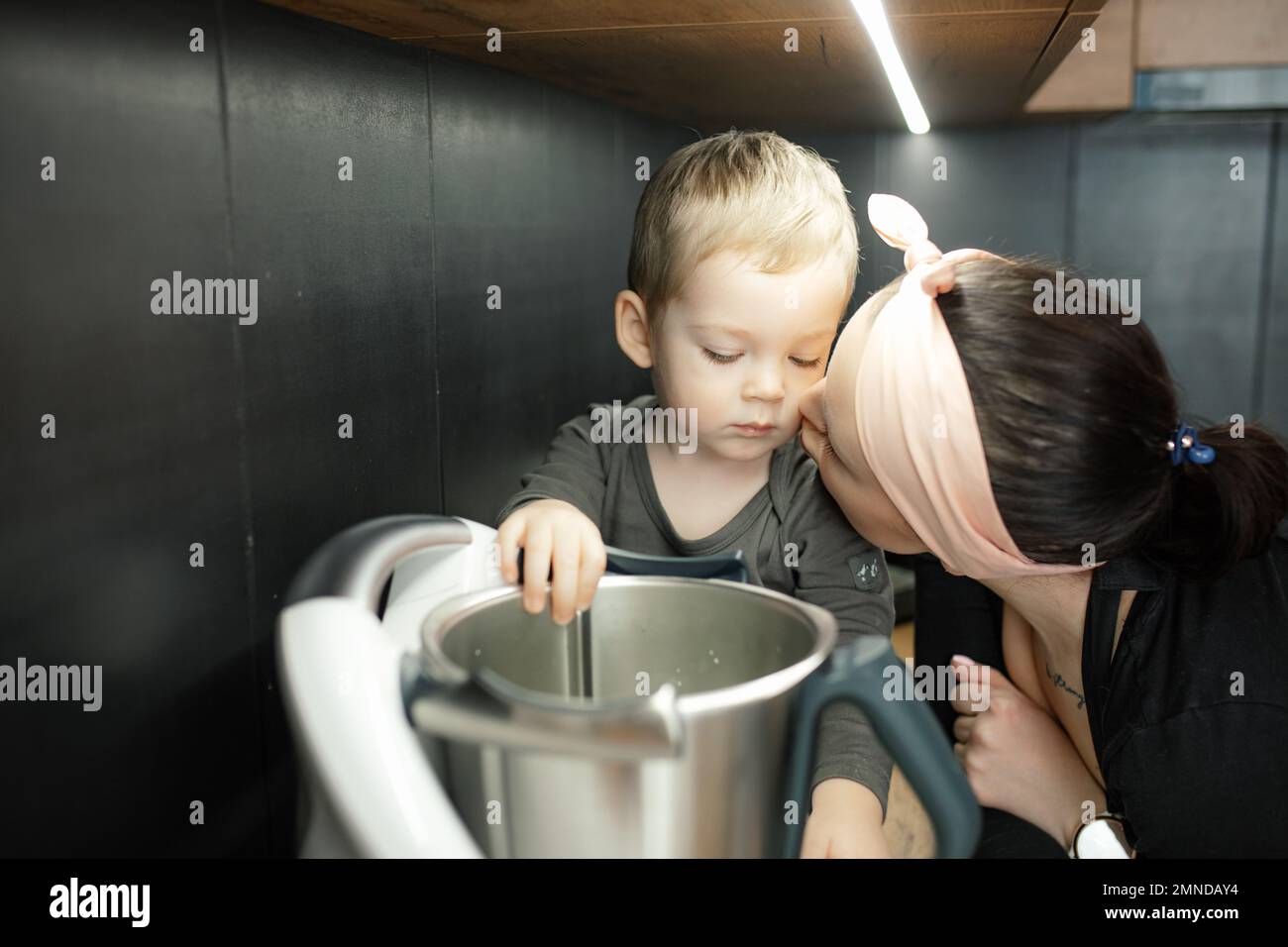 Mama mit Liebe küsst den kleinen Sohn auf die Wange. Baby, schau dir die tiefe Metallschale des Mixers an. Kleines Kind, hilf Mutter beim Kochen des Abendessens. Stockfoto