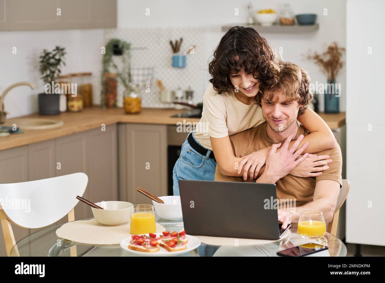 Glückliche junge Frau, die ihren Mann umarmt, während beide während der Kommunikation in einem Video-Chat nach dem Frühstück auf den Laptop-Bildschirm schauten Stockfoto