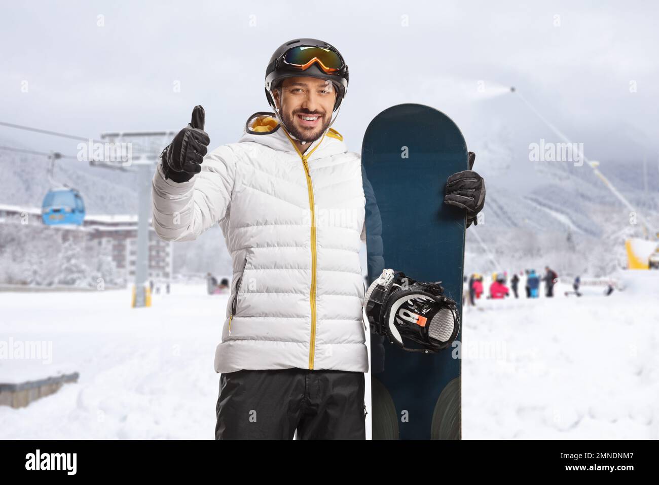Junger Mann mit Snowboard, Daumen hoch und lächelnd in einem Skigebiet Stockfoto