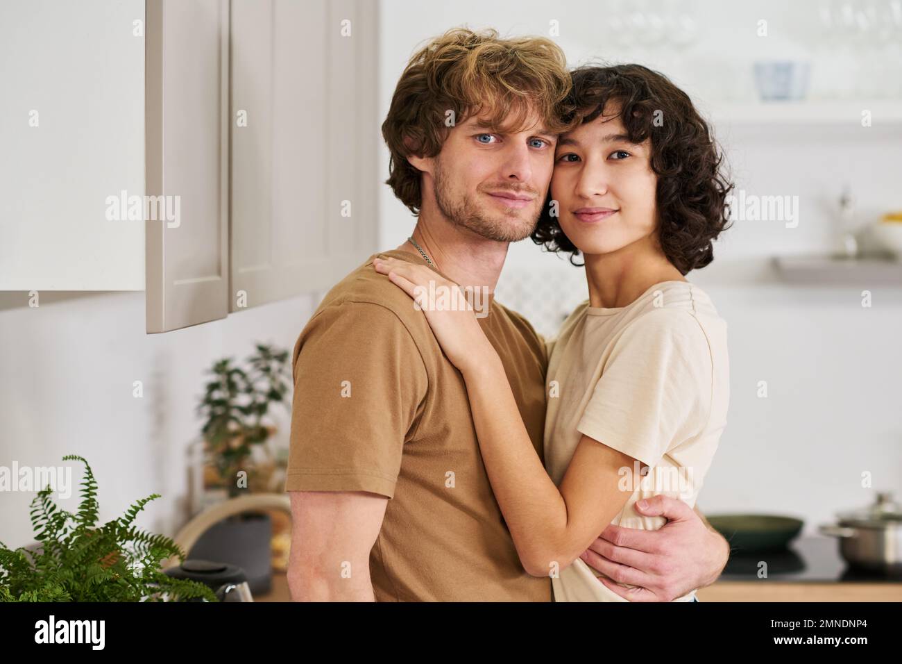 Ein junges, ruhiges, heterosexuelles Paar in T-Shirts, das sich an den Köpfen berührt, während es in der Küche in Umarmung steht und in die Kamera schaut Stockfoto