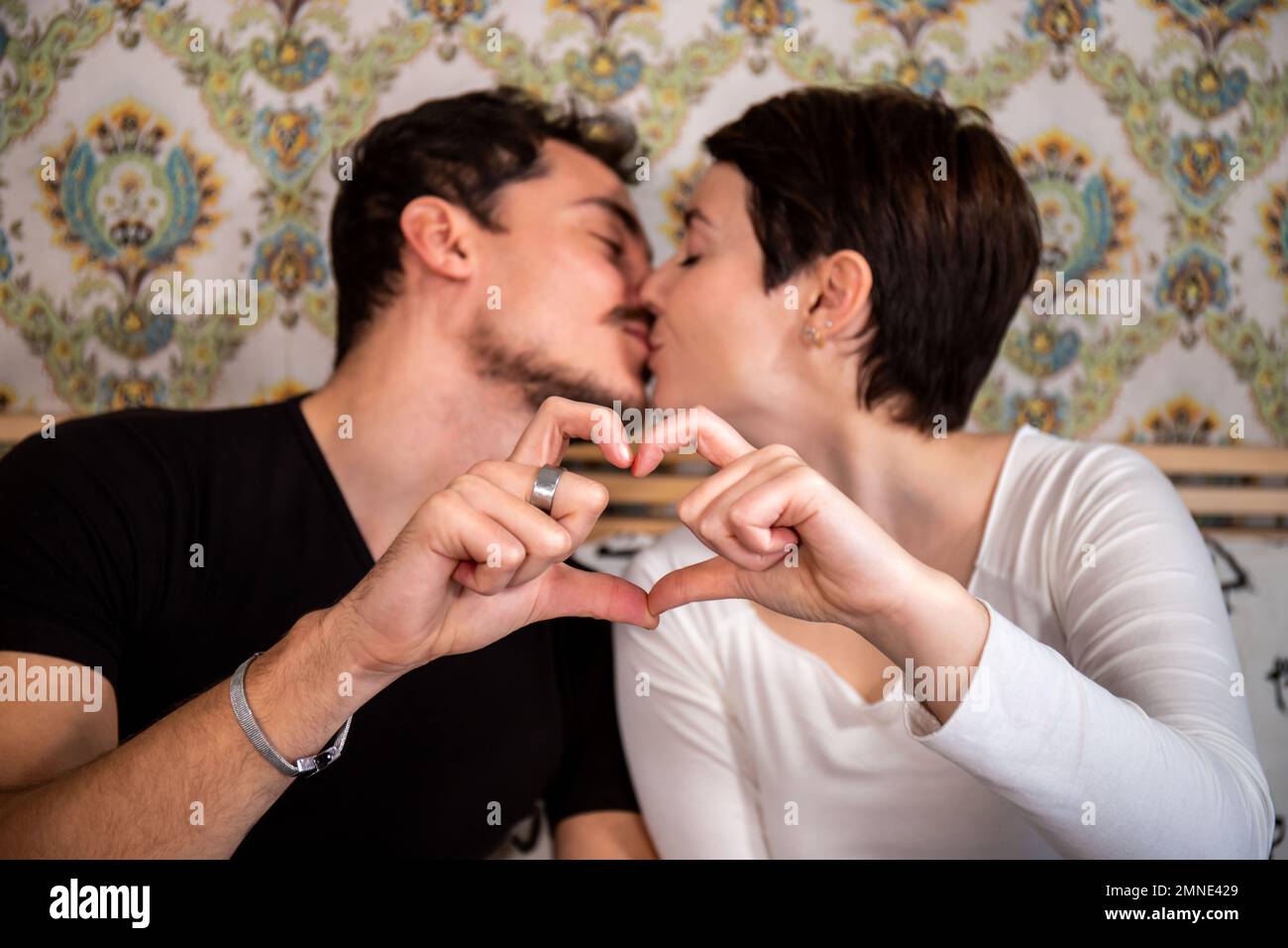 Das junge Paar sitzt auf dem Bett und küsst sich. Das Paar bildet mit seinen Händen ein Herz. Konzept von Liebe und Glück. Stockfoto