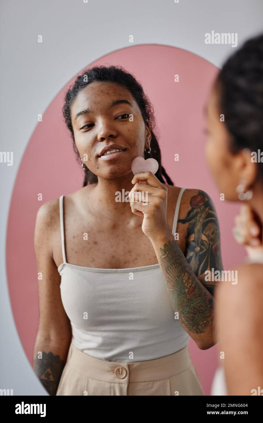 Vertikalporträt einer jungen schwarzen Frau mit Aknenarben unter Verwendung eines Massagegeräts während der Schönheitsroutine im Spiegel Stockfoto