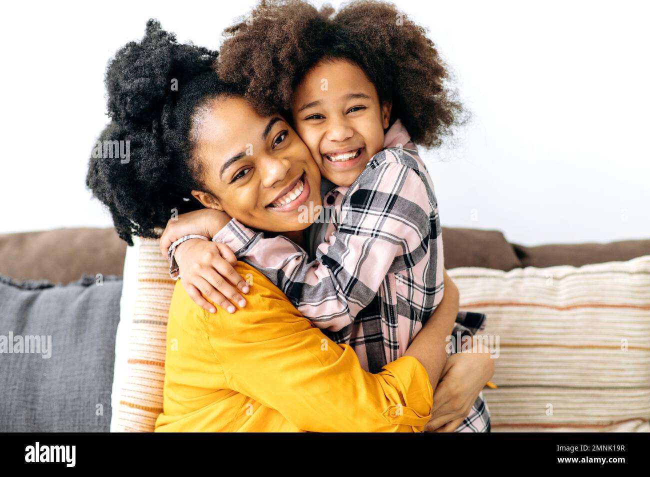 Fröhliche mutter und Kind. Afroamerikanische mutter und ihre süße Vorschultochter umarmen sich fest, während sie zu Hause auf dem Sofa sitzen, in die Kamera schauen, lächeln. Liebe zu Eltern und Kind Stockfoto