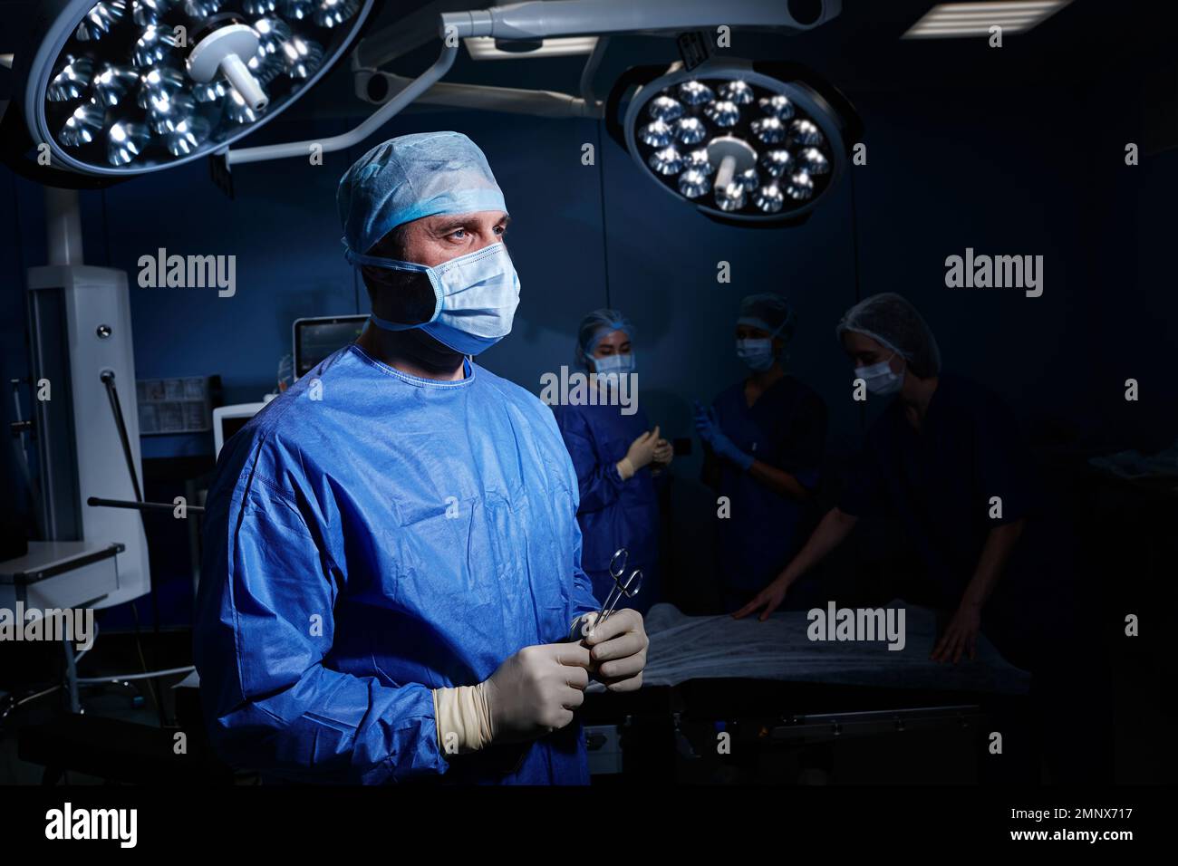 Beleuchtetes Porträt der männlichen Chirurgie mit PSA mit chirurgischer Schere in den Händen stehend im Operationssaal mit medizinischem Team Stockfoto