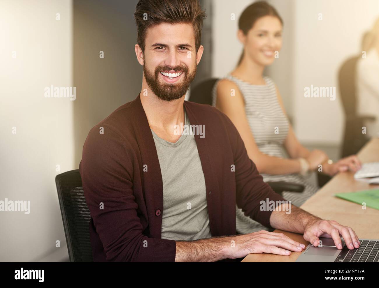 Erlauben Sie mir, mich um all Ihre Geschäftsanforderungen zu kümmern. Porträt eines jungen Designers, der in einem Büro sitzt, mit seinem Kollegen im Hintergrund verschwommen. Stockfoto