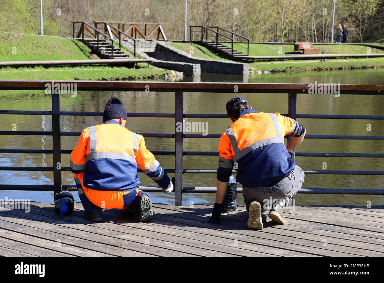 Arbeiter in Uniform streichen den Metallzaun an einem Seeufer. Bau- und Reparaturarbeiten im Frühlingspark, Vorbereitung auf die Strandsaison Stockfoto