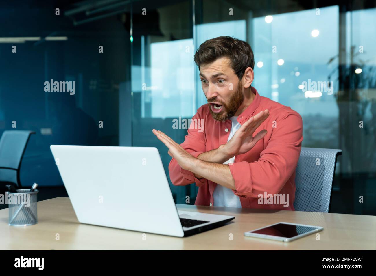 Probleme bei der Arbeit. Ein wütender junger Mann in einem roten Hemd sitzt im Büro und spricht über ein Videogespräch von einem Laptop aus. Schreie, aggressives Gesten mit seinen Händen, leugnet. Stockfoto