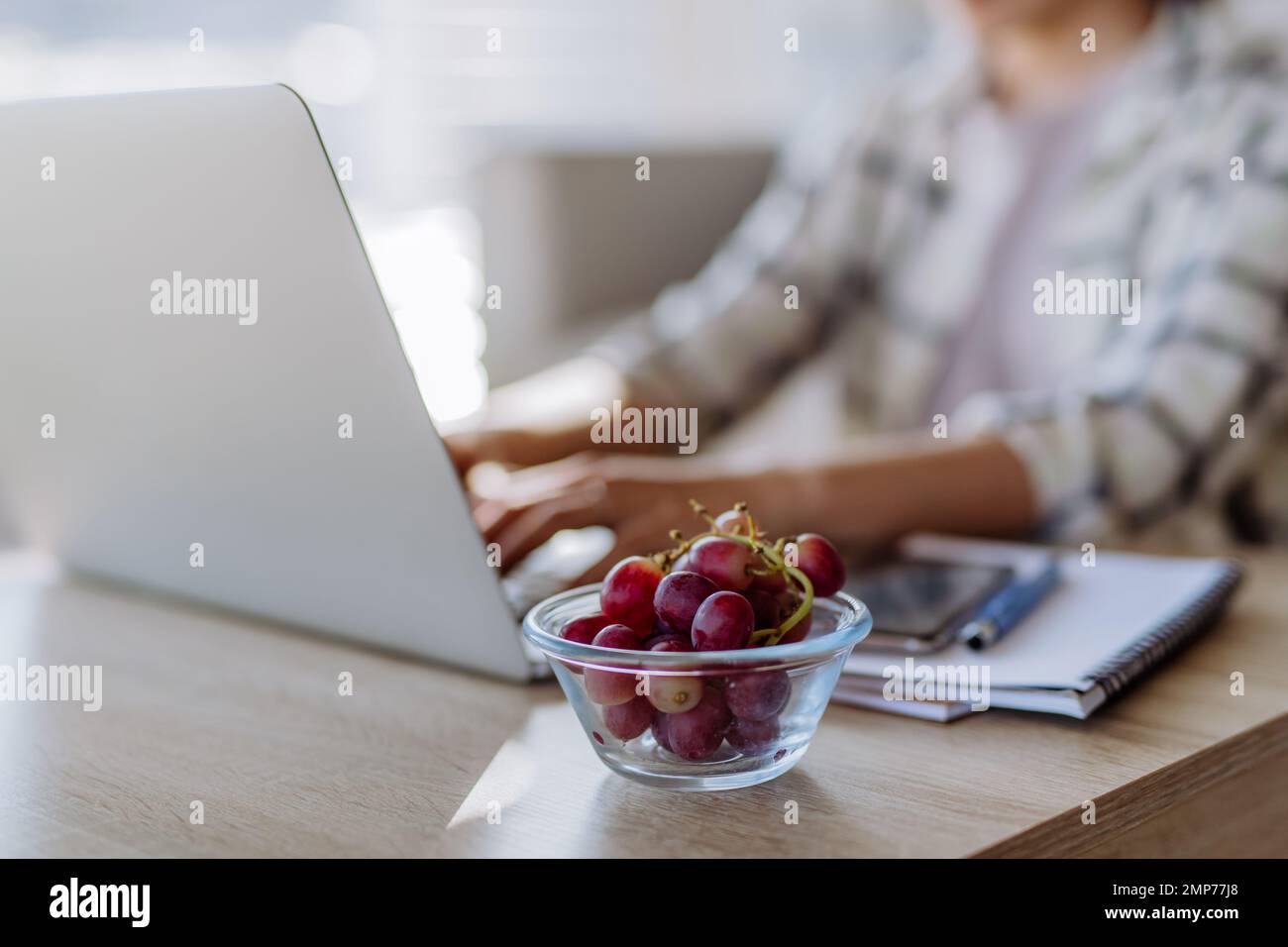 Seitenansicht einer Frau, die eine Schüssel mit Trauben über dem Schreibtisch hält, mit Computer, Tagebuch und Smartphone. Konzept der Work-Life-Balance. Stockfoto