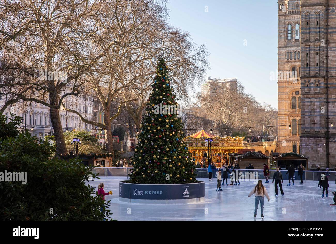 Die Eislaufbahn im Natural History Museum in London ist tagsüber geöffnet, mit einem Weihnachtsbaum in der Mitte und einem Karussell im Hintergrund. Stockfoto