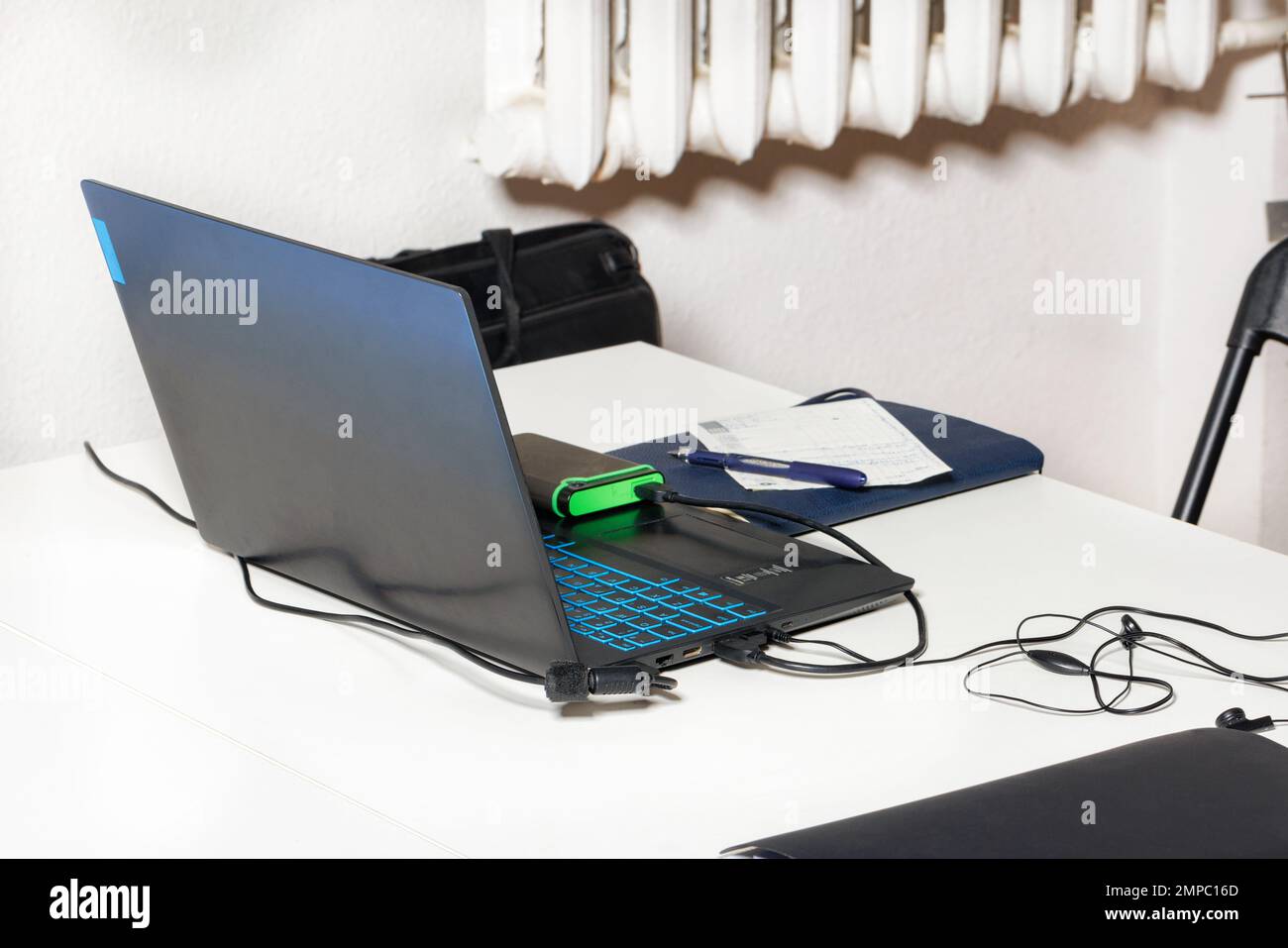 Laptop, Netzteil, Powerbank, Notizblock und Stift auf einem weiß laminierten Tisch mit einem unscharfen hellen Hintergrund. Seitenansicht. Moderne Geräte und Gadgets. Stockfoto