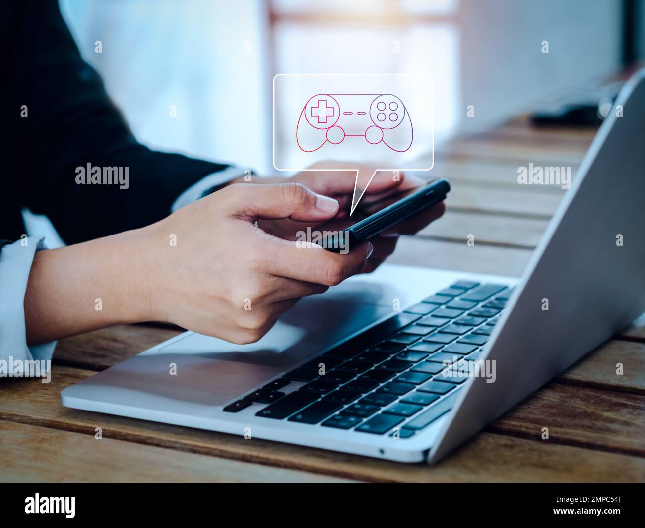 Spielcontroller, Joystick-Symbol in Sprechblase Symbol erscheint auf dem Smartphone in den Händen des Geschäftsmanns, horizontal, während ein Notebook arbeitet Stockfoto