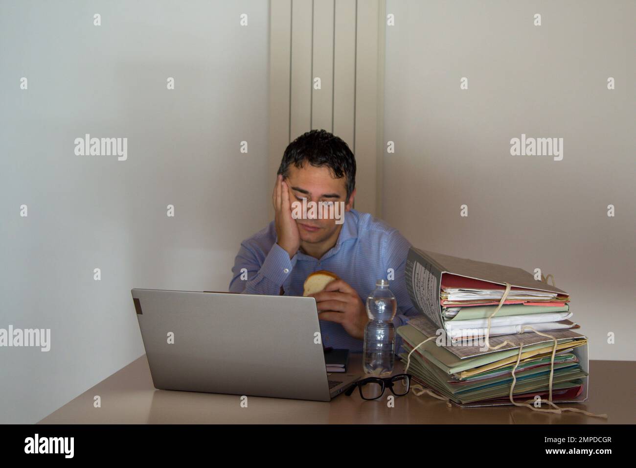 Bild eines verzweifelten Mannes mit einem Sandwich und einer kleinen Flasche Wasser, der wegen Überstunden in seinem Büro arbeiten muss. Stockfoto