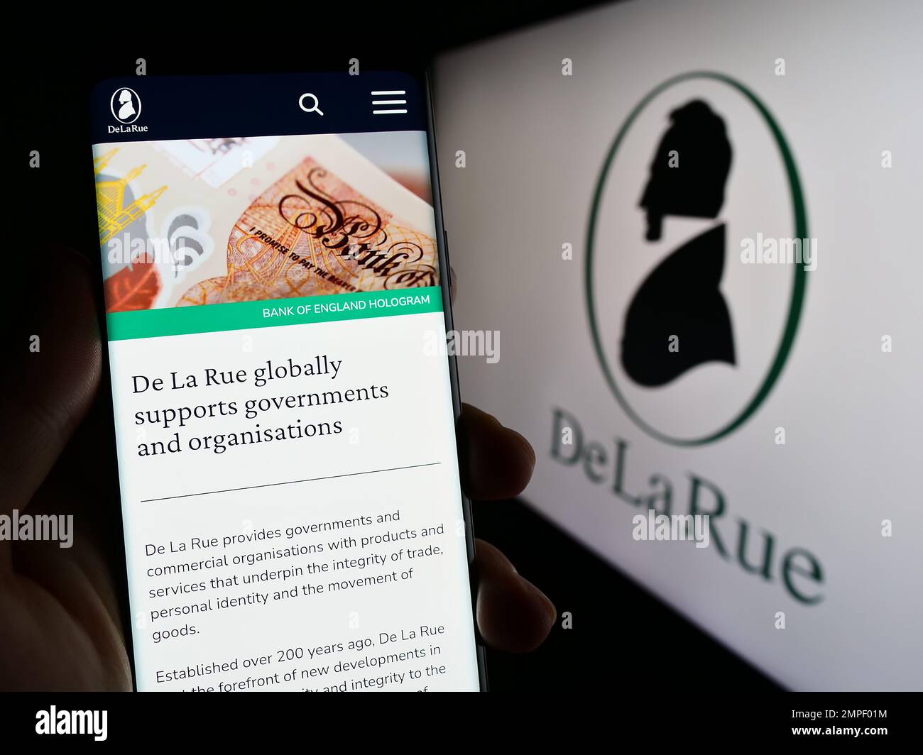 Person, die ein Mobiltelefon mit einer Webseite des britischen Banknotenproduzenten De La Rue plc auf dem Bildschirm mit Firmenlogo hält. Konzentrieren Sie sich auf die Mitte des Telefondisplays. Stockfoto
