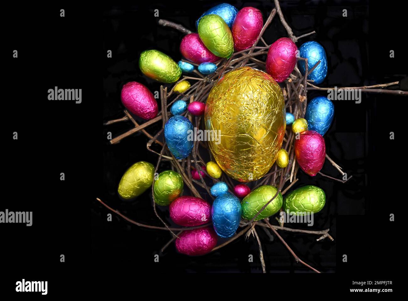 In Folie verpackte mehrfarbige ostereier in rosa, grün, blau und gelb in einem natürlichen Nest aus Holz, Stäbchen und Zweigen, vor schwarzem Hintergrund. Stockfoto