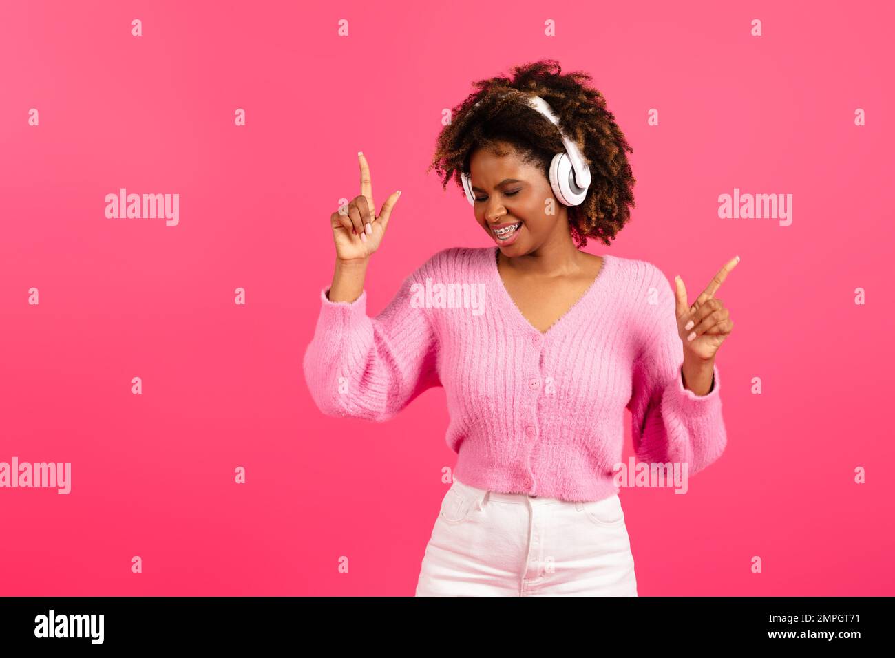 Fröhliche junge schwarze lockige Frau in legeren, kabellosen Kopfhörern mit Zahnspangen, die mit den Fingern nach oben tanzen Stockfoto