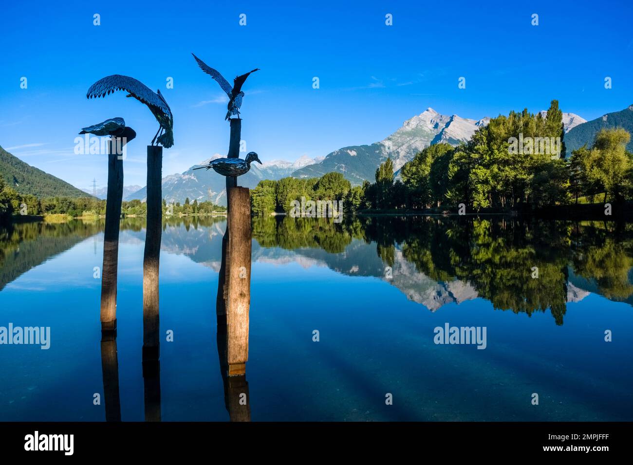 Kunstinstallation mit Metallvögeln auf Stangen in einem kleinen See in der Nähe des Flughafens von Sion, alpine Landschaft in der Ferne. Stockfoto
