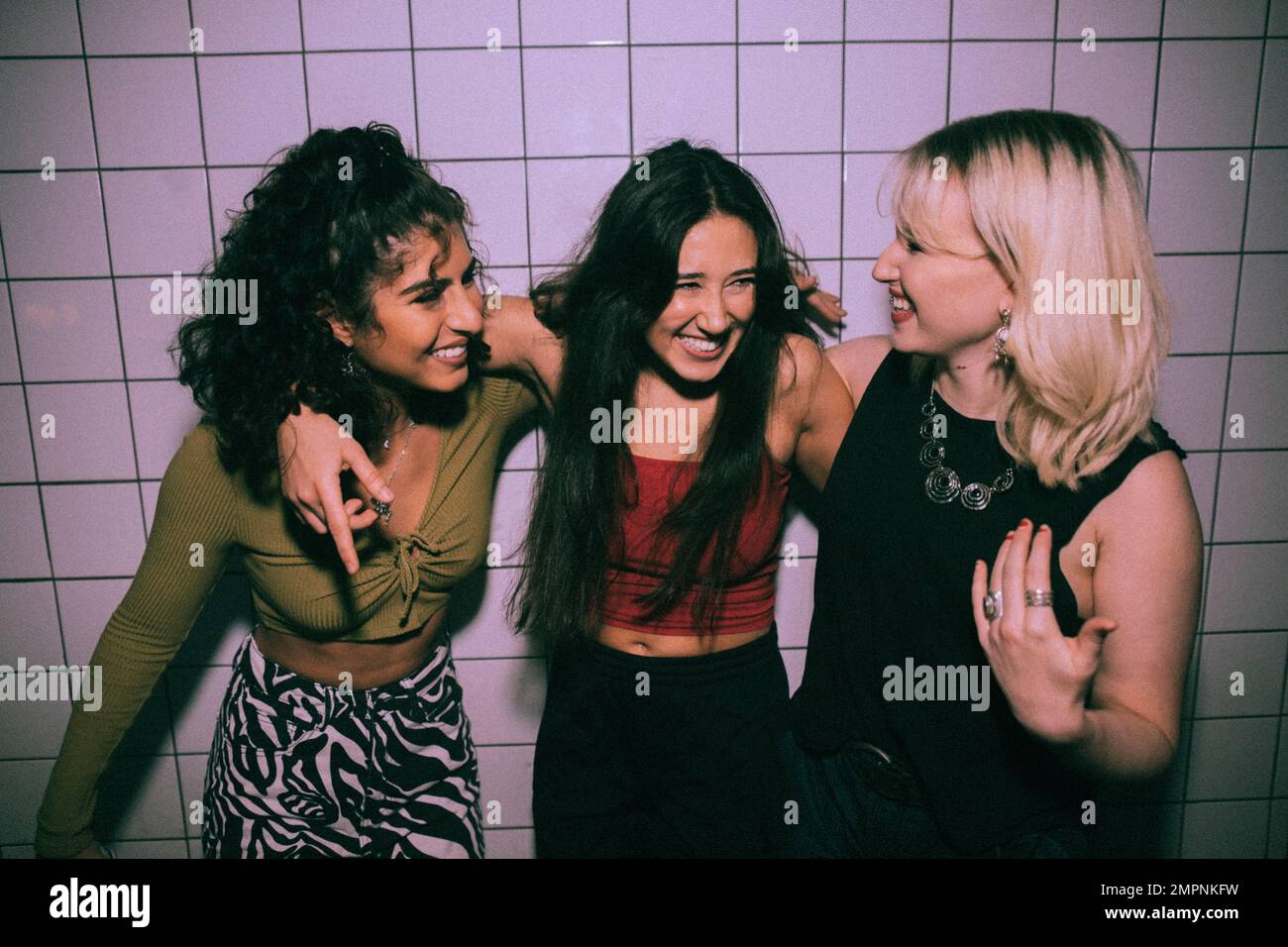 Fröhliche, junge, multirassische, weibliche Freunde, die mit Armen gegen die weiße Wand im Nachtclub stehen Stockfoto