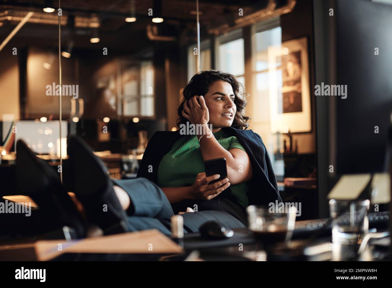 Geschäftsfrau mit der Hand im Haar, die ein Smartphone hält und mit erhobenen Füßen am Schreibtisch im Büro sitzt Stockfoto