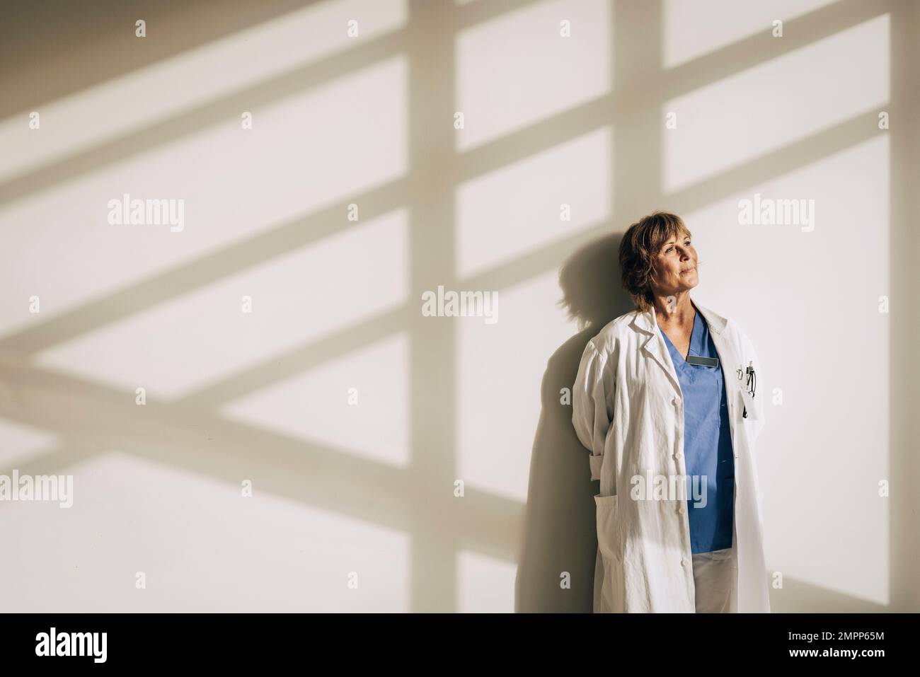 Eine Ärztin trägt einen Laborkittel und träumt, während sie mit Sonnenlicht an der Wand steht Stockfoto