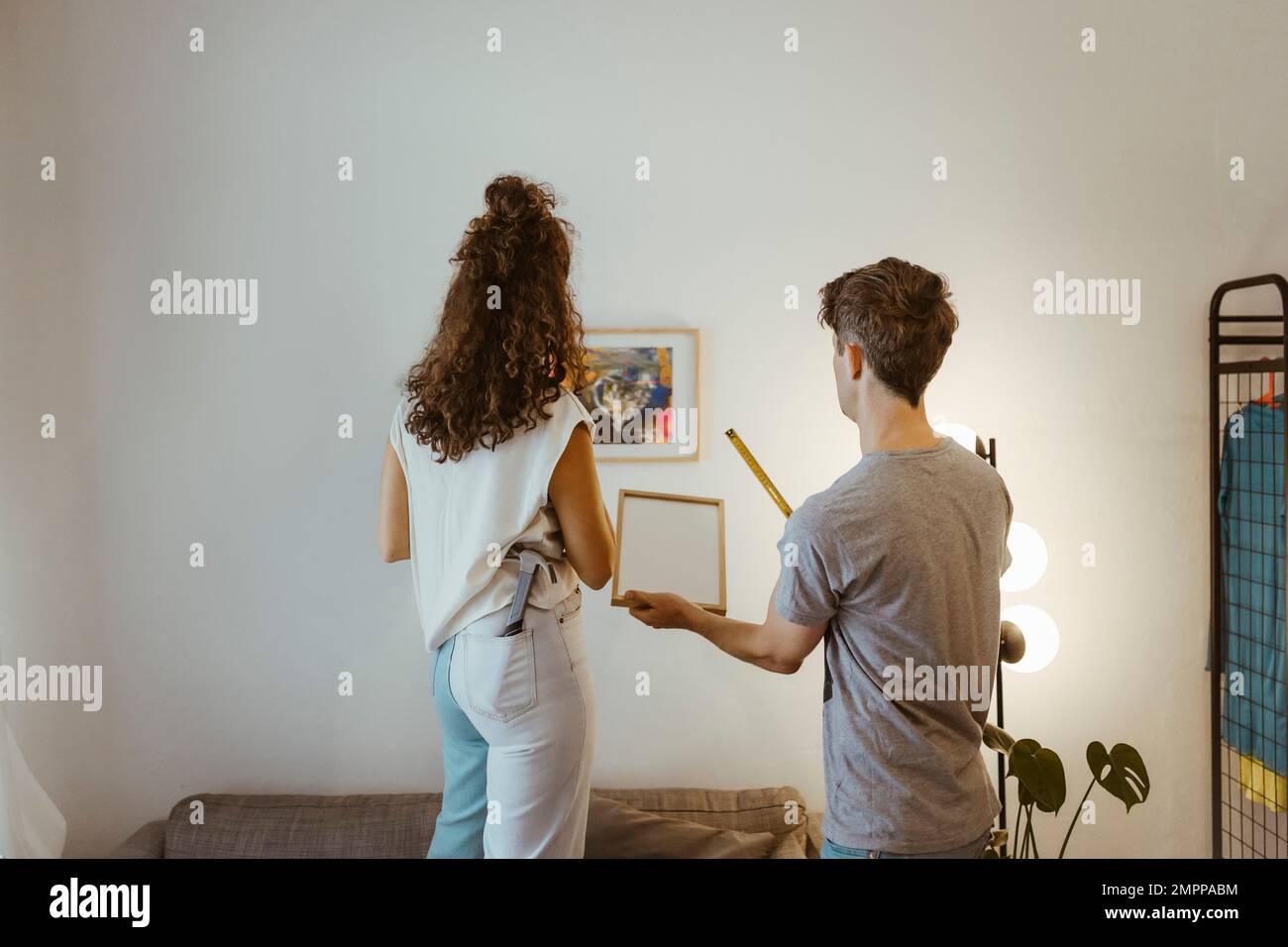 Rückansicht von Freund und Freundin, die sich gegenseitig helfen, während der Rahmen zu Hause an der Wand montiert wird Stockfoto