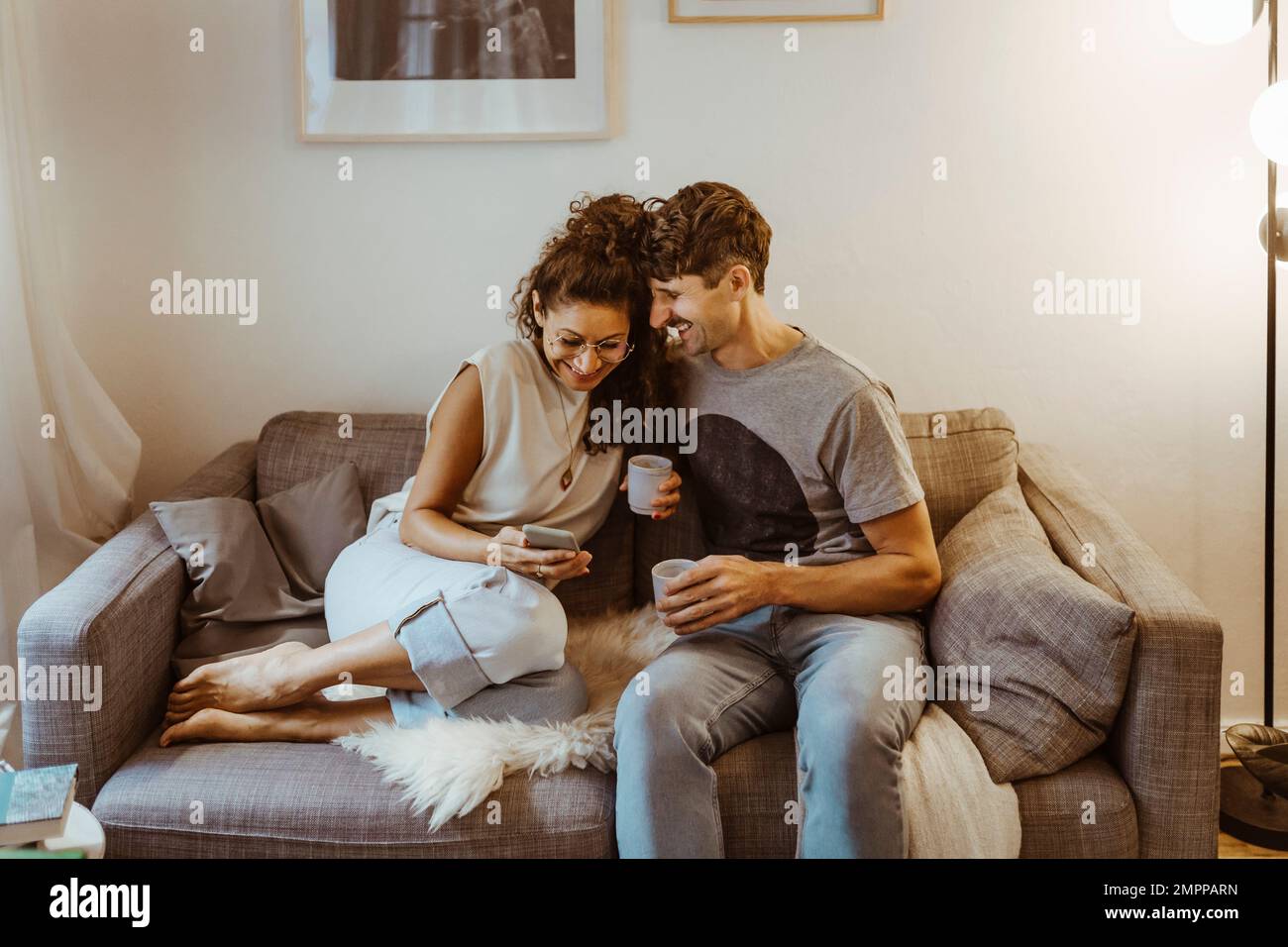 Glückliches Paar, das Kaffee trinkt, während es sein Smartphone mit anderen teilt und zu Hause auf dem Sofa sitzt Stockfoto