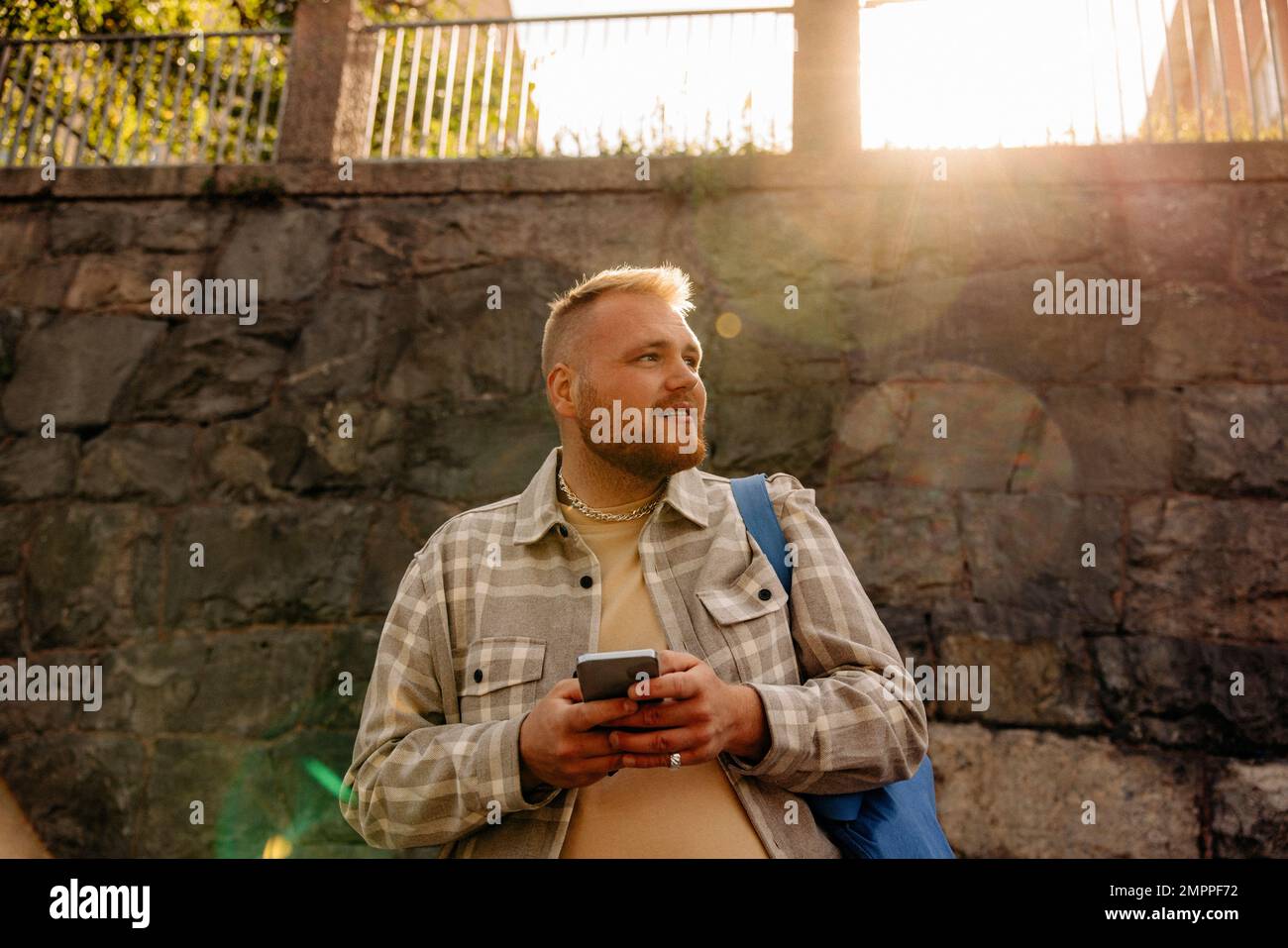 Ein Mann, der an sonnigen Tagen vor der Wand steht und sein Smartphone in der Hand hält und wegschaut Stockfoto