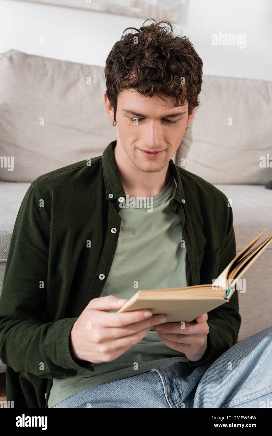 Intelligenter Mann mit lockigem Haar liest Buch im Wohnzimmer, Stockbild Stockfoto