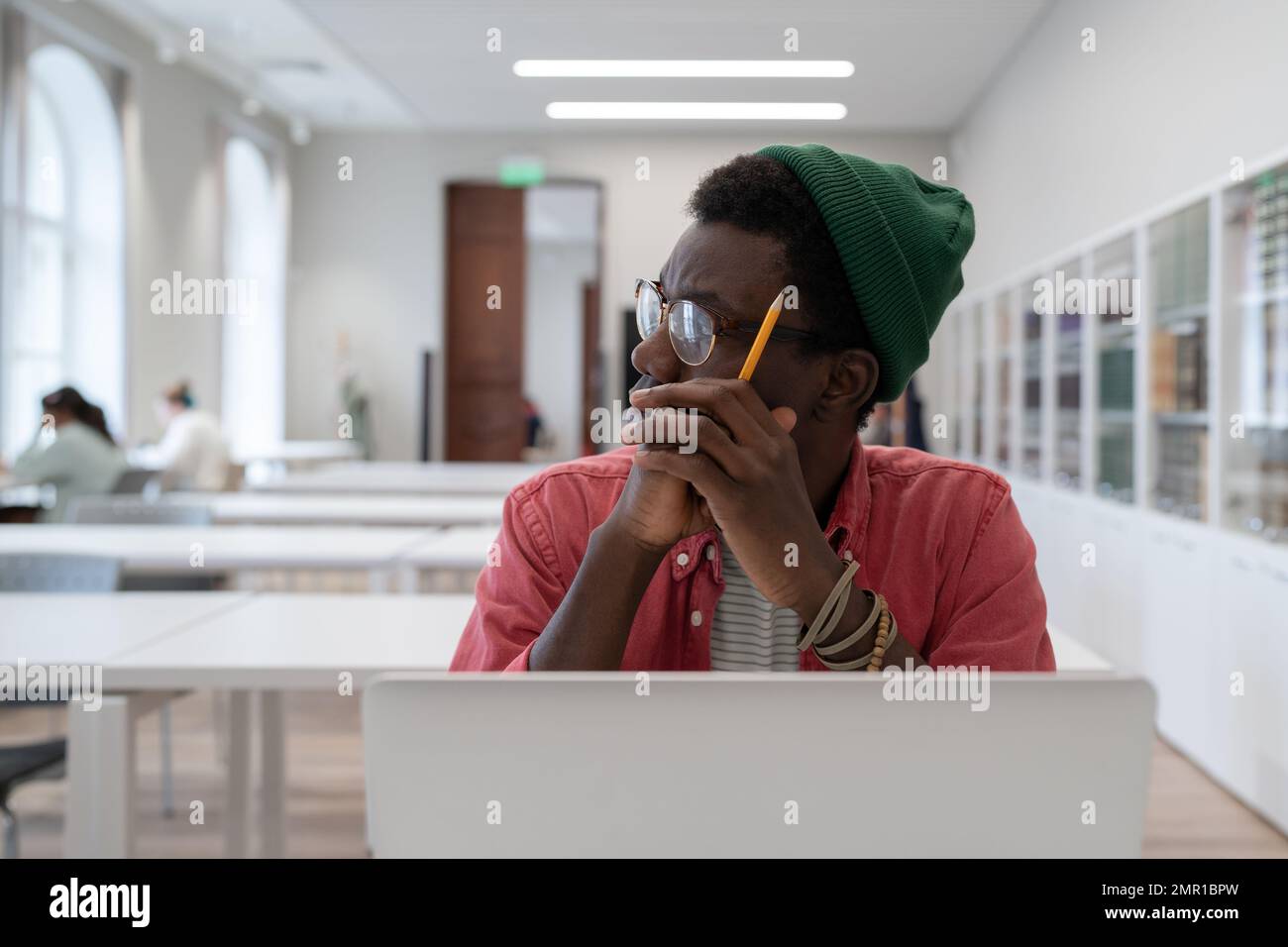 Ein afrikanischer Student, der vom Lernen abgelenkt war, am Tisch mit einem Laptop in der Bibliothek saß und zur Seite schaute Stockfoto