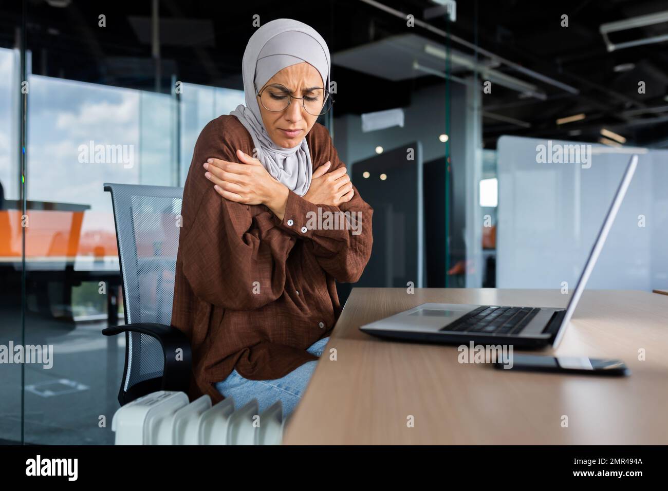 Eiskalte arabische Frau im Büro, Geschäftsfrau im Hijab-Kaltluftheizung und Heizung funktionierten nicht, benutzte Laptop am Arbeitsplatz, versuchte zu wärmen. Stockfoto