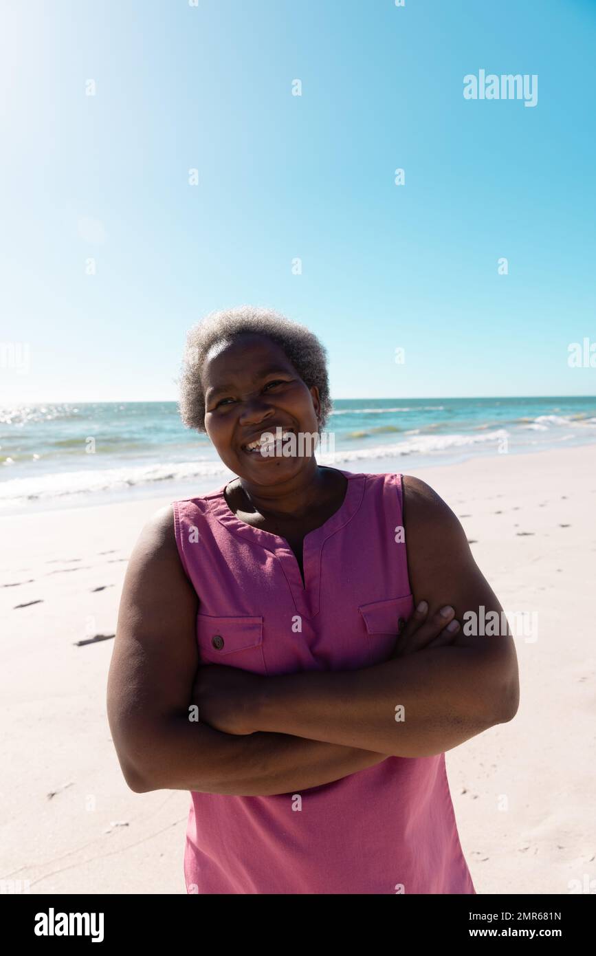 Fröhliche afroamerikanische Seniorin mit gekreuzten Armen, die vor dem malerischen Blick auf das Meer und den Himmel steht Stockfoto