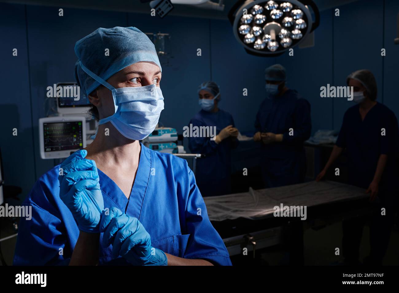 Beleuchtetes Porträt einer chirurgischen Krankenschwester, die sterile Handschuhe vor der Operation legt, während sie auf der Station mit Chirurgen und Assistenten steht Stockfoto