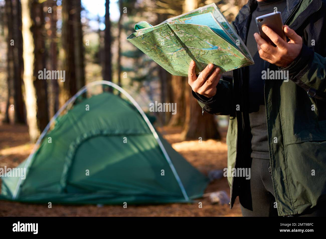 Erwachsener Wanderer mit Rucksack, der Handy und Karte benutzt, im Wald in der Nähe seines Zelts. Wandern älterer Mann in der herbstlichen Natur mit Karte im Freien neben einem Stockfoto