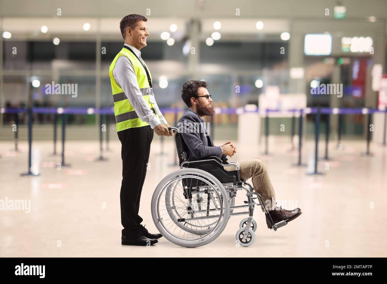 Profilaufnahme über die gesamte Länge eines Sonderhelfers am Flughafen mit einem männlichen Passagier im Rollstuhl in einem Terminalgebäude Stockfoto